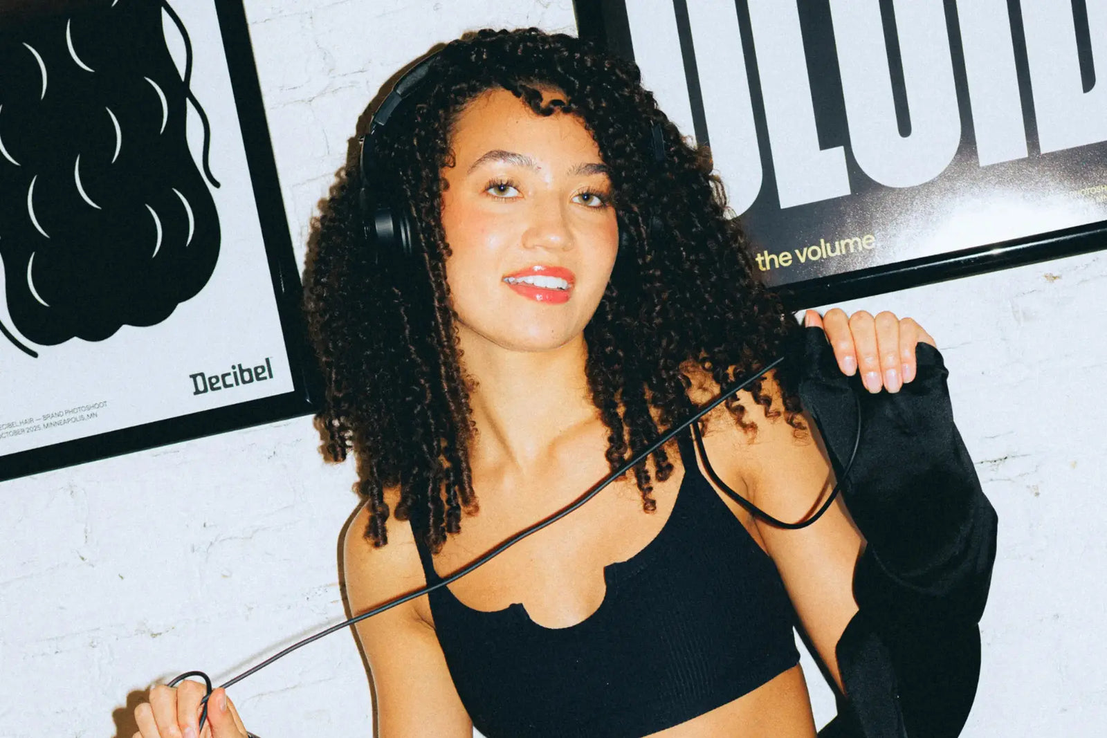 Curly haircare model with voluminous natural curls and headphones, seated on a tan leather couch in front of Decibel Hair brand posters.