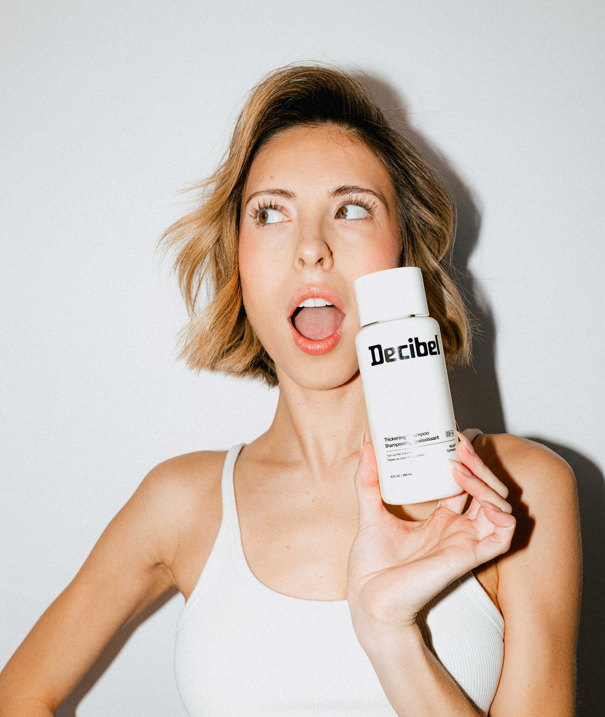 Lifestyle photo of woman with voluminous hair holding Decibel Thickening Shampoo bottle with an open-mouth expression.