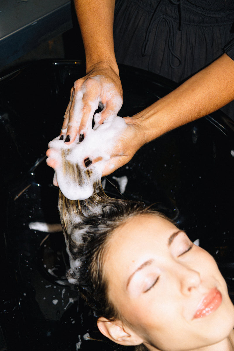 Step 1: Stylist lathering Decibel Thickening Shampoo through a client's hair at a salon wash basin.