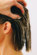 Hand massaging creamy thickening conditioner into wet brunette hair near the scalp.