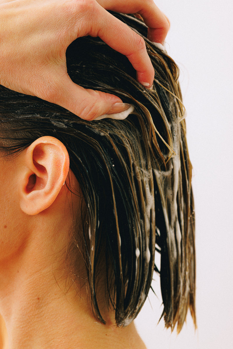 Hand massaging creamy thickening conditioner into wet brunette hair near the scalp.